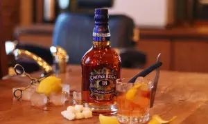 Butelka Chivas Regal 18, szklanka z drinkiem, kostki lodu, cytryna i cukier na drewnianym stole.