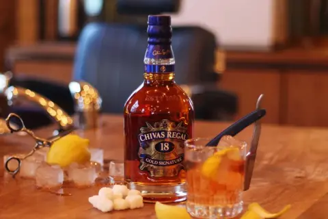 Butelka Chivas Regal 18, szklanka z drinkiem, kostki lodu, cytryna i cukier na drewnianym stole.