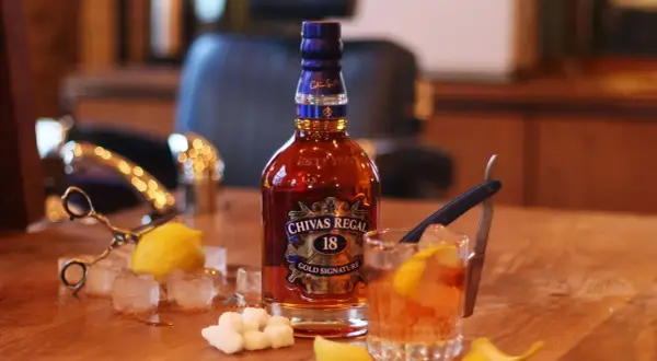 Butelka Chivas Regal 18, szklanka z drinkiem, kostki lodu, cytryna i cukier na drewnianym stole.